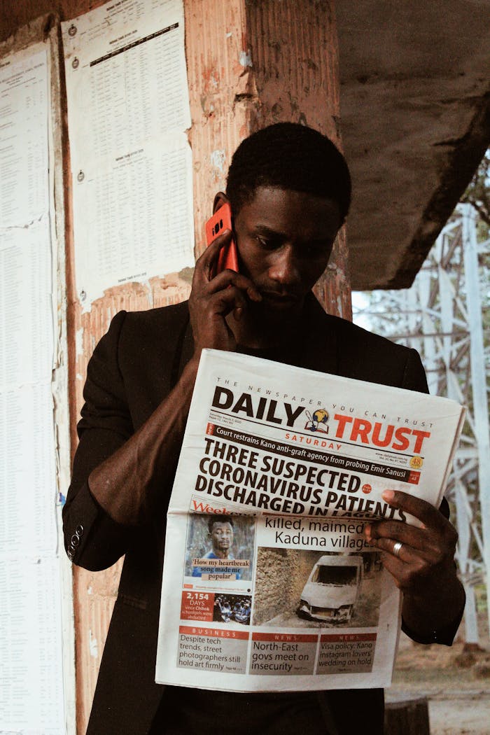 Serious African American male with short hair standing near shabby bus station with newspaper and talking by smartphone